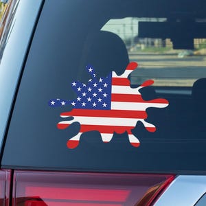 May include: A car window decal featuring a stylized American flag design. The flag is depicted in a splash-like shape with red and white stripes and a blue field of stars. The decal is applied to the rear window of a vehicle.