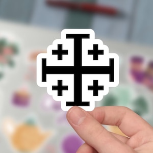 Puede incluir: Una pegatina en blanco y negro con el diseño de la cruz de Jerusalén. La cruz tiene un contorno negro grueso y está rellena de negro. La pegatina está sujeta en la mano de una persona, con un fondo borroso de un portátil y otros objetos.