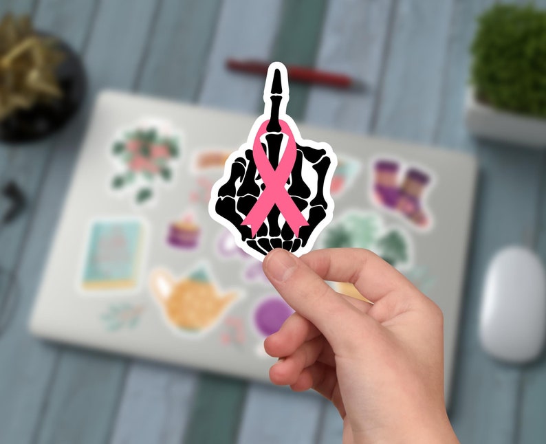 May include: A black and white sticker featuring a skeletal hand giving the middle finger, with a pink ribbon tied around the middle finger. The sticker is held in a person's hand, with a blurred background of other stickers and a laptop.