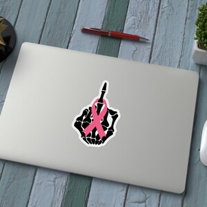 May include: A laptop with a sticker featuring a black skeleton hand giving the middle finger, adorned with a pink ribbon. The image is on a wooden surface with a pen, plant, and mouse.