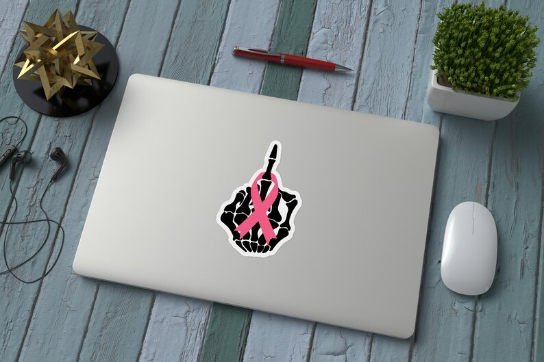 May include: A laptop with a sticker featuring a black skeleton hand giving the middle finger, adorned with a pink ribbon. The image is on a wooden surface with a pen, plant, and mouse.