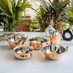 Op de afbeelding: Een set van vier gehamerde koperen serveerschalen en een koperen emmer, elk met een gepolijste zilverkleurige binnenkant. De schalen hebben twee zijhandgrepen, terwijl de emmer een zwenkhandgreep heeft. Elk stuk heeft een klein gegraveerd logo.