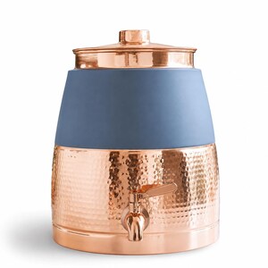 Puede incluir: Un dispensador de bebidas con una base de cobre martillado y una sección superior de cerámica azul pizarra mate. La unidad incluye una tapa de cobre a juego y un grifo de cobre funcional centrado en la parte frontal para servir líquidos.