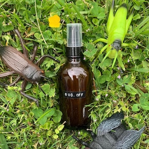 May include: A brown glass bottle with a black label that says "BUG OFF" on it. The bottle is surrounded by fake insects, including a green grasshopper, a brown cricket, and a black fly.