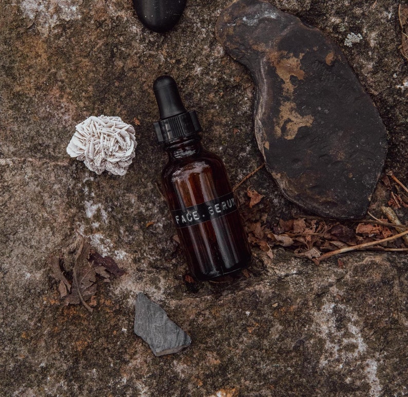May include: A dark brown glass bottle with a dropper top and a black label that reads "FACE SERUM". The bottle is sitting on a rocky surface with a small white rock and a piece of slate.