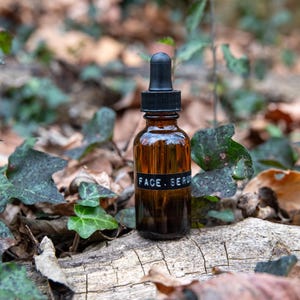 May include: A brown glass bottle with a black dropper top and a black label that reads "FACE. SERUM". The bottle is sitting on a piece of wood with green leaves and brown leaves around it.