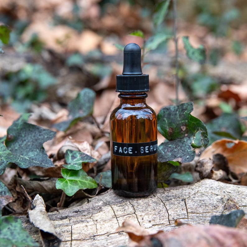 May include: A brown glass bottle with a black dropper top and a black label that reads "FACE. SERUM". The bottle is sitting on a piece of wood with green leaves and brown leaves around it.