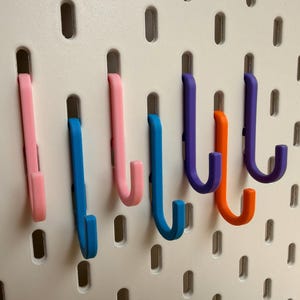 May include: A set of six J-shaped hooks in pink, blue, orange, and purple colors, inserted into a white pegboard with vertical slots. The hooks are arranged in a row, each featuring a curved end for hanging items, made of smooth plastic material.