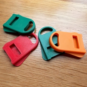 May include: Four plastic bag handle carriers in green, red, and orange. Each carrier features a rounded handle opening and a rectangular slot for securing bag straps. The items have a textured surface finish and are arranged in a small cluster.