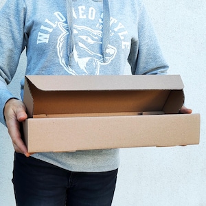 Puede incluir: Una persona sosteniendo una caja de cartón rectangular abierta. La caja es de cartón corrugado y tiene un color marrón natural. La persona lleva una sudadera con capucha gris con el texto "WILD STYLE" y un gráfico.