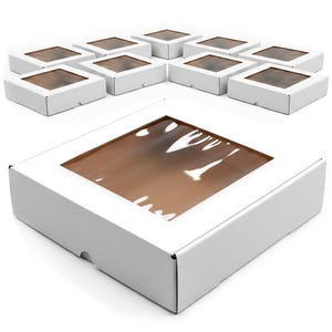 Op de afbeelding: Een verzameling witte kartonnen dozen met transparante, vierkante vensters. De dozen zijn gegroepeerd, met één doos op de voorgrond. De vensters tonen een bruin interieur, wat suggereert dat de dozen bedoeld zijn voor het presenteren of verpakken van items.