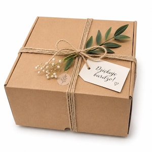 Può includere: Una scatola regalo quadrata in cartone marrone legata con spago di juta naturale. La scatola è decorata con un piccolo rametto di foglie verdi, un grappolo di fiori bianchi e un'etichetta bianca con il testo Dziękuję bardzo! e un cuore.