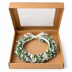 May include: A floral crown of white flowers and greenery, displayed in an open cardboard box with a clear window in the lid. The crown is tied with a white ribbon. The box is brown.