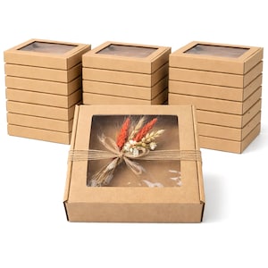 Pode incluir: Caixas de presente cor kraft com tampas transparentes. Uma caixa é decorada com um buquê de flores secas e amarrada com barbante. Pilhas de caixas são dispostas ao fundo.