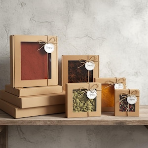 May include: A selection of spice boxes with clear windows, tied with twine and labelled. The boxes contain various spices, including a red powder, star anise, green cardamom, yellow turmeric, and peppercorns. The boxes are on a wooden shelf.