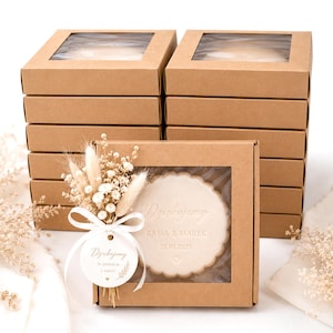 Op de afbeelding: Een stapel bruine kartonnen dozen met transparante deksels. Eén doos toont een ronde, lichtgekleurde koek met de tekst "Dziękujemy", namen en een datum. Een klein boeket droogbloemen en een label zijn bevestigd.
