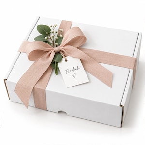 May include: A rectangular white gift box wrapped with a dusty pink ribbon tied in a bow. A small white gift tag with the text "Für dich" and a heart symbol is attached to the ribbon, along with a sprig of green eucalyptus and white baby's breath.