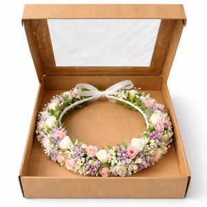 Op de afbeelding: Een bloemenkrans met witte, roze en paarse bloemen en groene bladeren, vastgebonden met een wit lint, gepresenteerd in een bruine kartonnen doos met een doorzichtig venster.