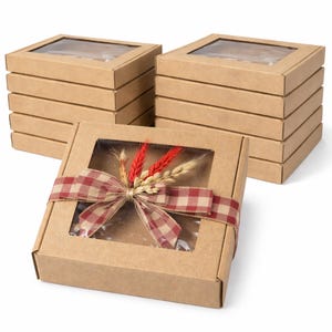 Op de afbeelding: Verschillende gestapelde, vierkante kartonnen dozen met transparante deksels. Eén doos is versierd met een rood-wit geruit lint en een bundel tarwe en rode decoratieve stelen. De dozen zijn gerangschikt op een witte achtergrond.