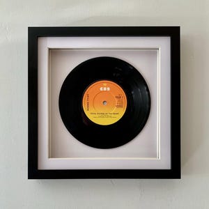 May include: Framed vinyl record of Bonnie Tyler's "Total Eclipse of the Heart." The black vinyl record is centered in a white mount, encased in a black square frame. The record label is orange and yellow with black text. The frame is mounted on a light grey wall.