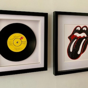 May include: Two framed pieces of art. The left frame contains a black vinyl record with a yellow label that reads "The Rolling Stones -  'Shattered'  (UK 45 RPM). The right frame contains a black and white image of the Rolling Stones' iconic tongue logo with a red outline.
