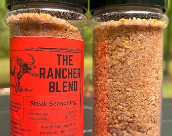 Puede incluir: Dos frascos de especias de plástico transparente con tapas negras llenos de un condimento para carne de color naranja parduzco. El frasco izquierdo tiene una etiqueta roja con una ilustración de un toro negro y el texto THE RANCHER BLEND, 198g.
