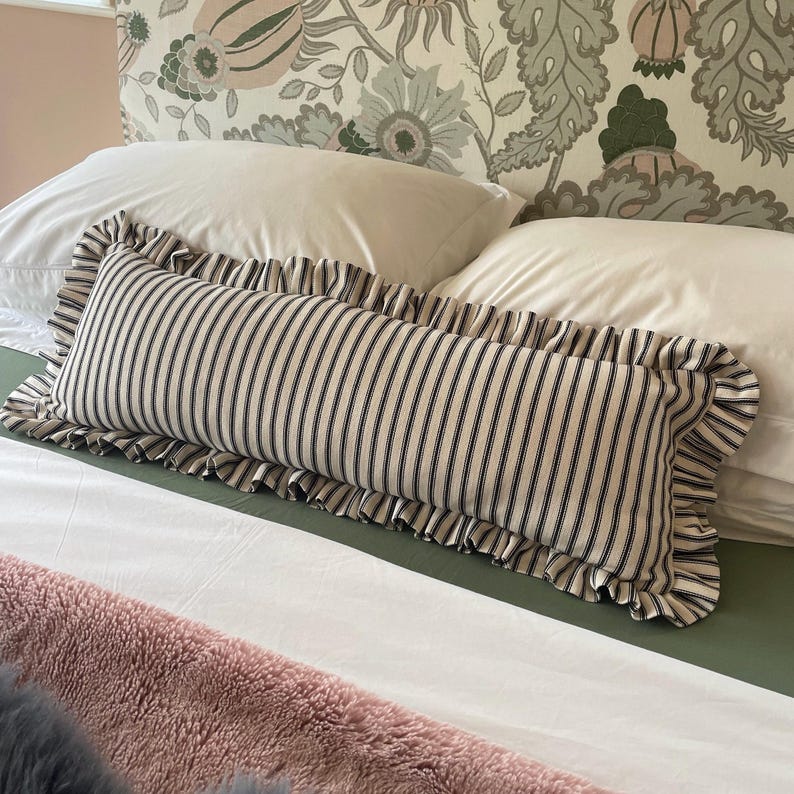 A white bed with a black and white striped pillow with frills. The headboard is covered in a floral fabric with green and pink flowers. A pink fluffy blanket is on the bed.