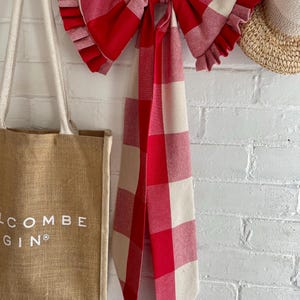 May include: A large fabric bow featuring a red and cream buffalo check pattern with ruffled edges and long hanging tails. The bow is displayed alongside a tan jute tote bag with the text SALCOMBE GIN printed in white and a straw hat.