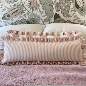 May include: A long, rectangular cushion with a pink and white striped design, edged with frills. It sits on a pink, fluffy blanket, with floral wallpaper behind. The cushion is designed for decorative use, and is likely for a bed.