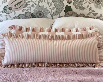 May include: A rectangular bolster pillow with a pink and white striped pattern, framed by ruffled edges. The pillow rests on a pink, textured blanket, with a floral patterned wallpaper in the background. The pillow is designed for decorative use.