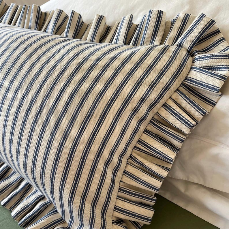 A white and blue striped pillow with a ruffled edge. The pillow is made of a soft, textured fabric and has a classic, farmhouse style.