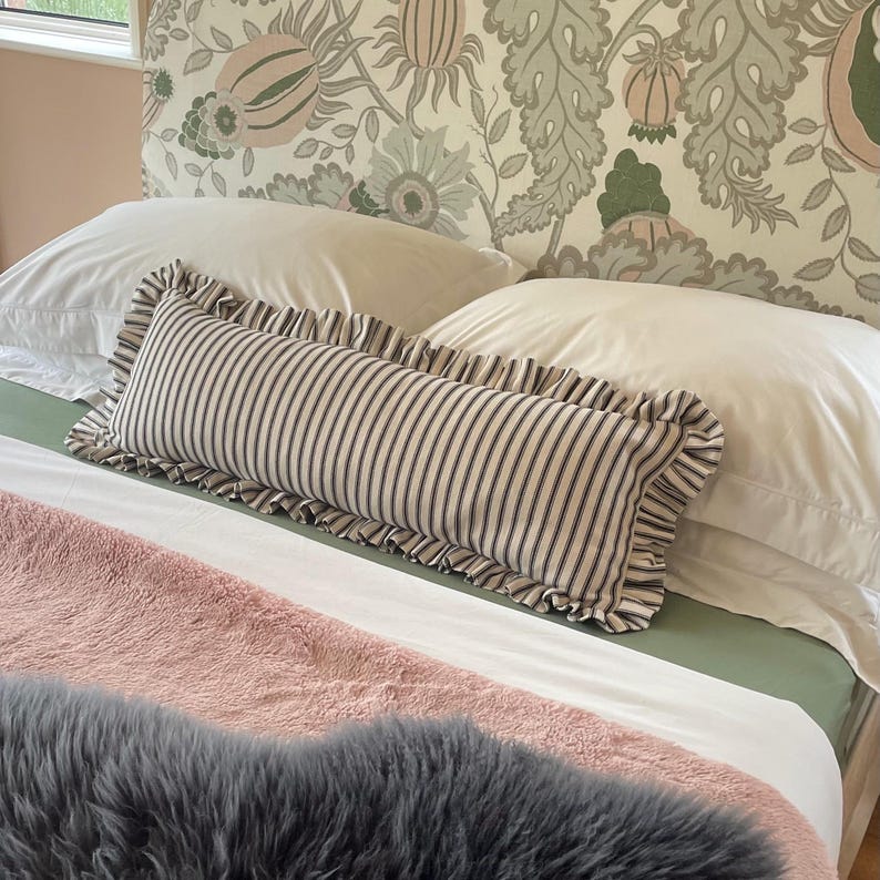 A white bed with a black and white striped pillow with frills. The headboard is covered in a floral fabric with green and pink flowers. A pink fluffy blanket is on the bed.