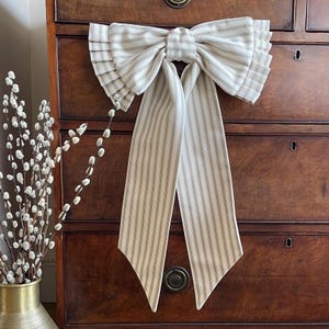 May include: A large decorative fabric bow featuring a cream and brown pinstripe pattern. The bow has pleated side edges and two long, vertical hanging tails that taper to pointed ends. The fabric is structured with a central knot detail.