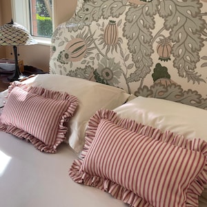 May include: Two rectangular cushions with red and white stripes and frilled edges on a white bed. A floral patterned headboard is behind the cushions. A lamp with a stained glass shade sits on a bedside table.