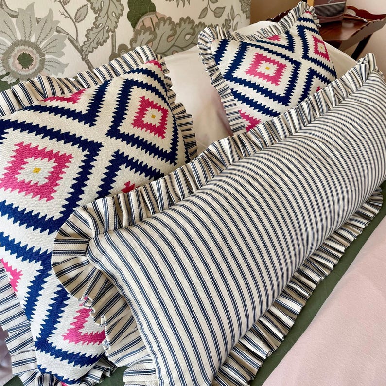 Three decorative pillows with a white background and blue and pink geometric patterns. The pillows have a white ruffle trim. The pillows are on a bed with a floral patterned headboard.