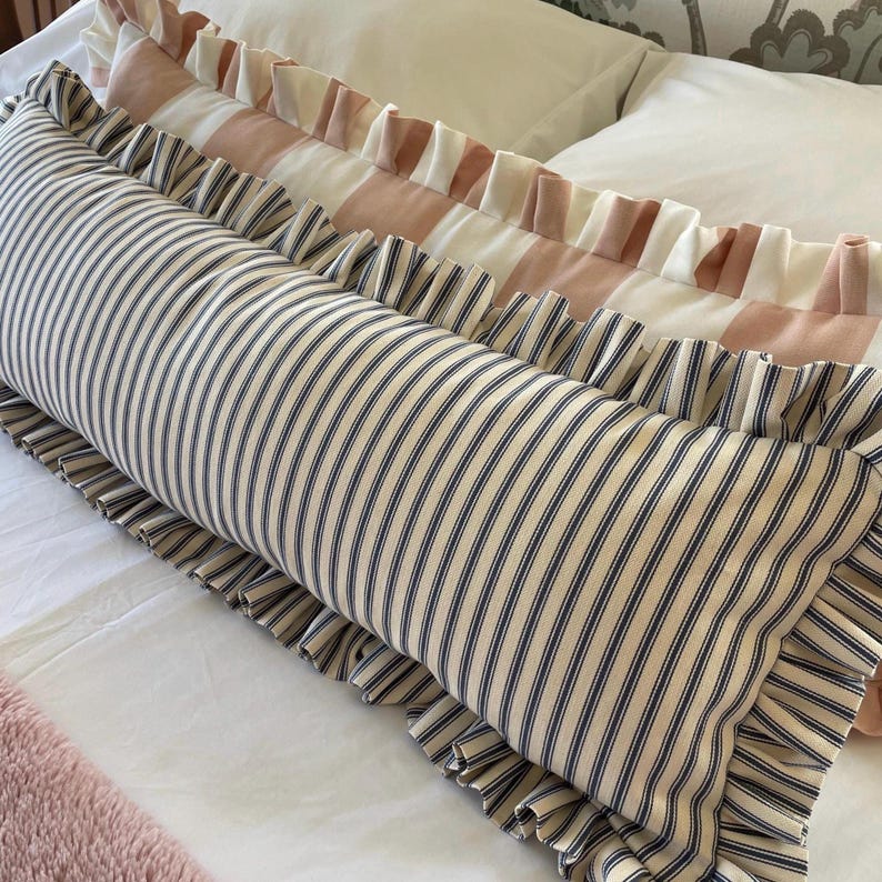 A striped lumbar pillow with ruffled trim in navy blue cotton ticking on a bed with a pink bedspread.