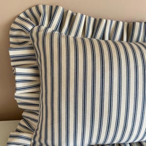A white and blue striped pillow with a ruffled edge. The pillow is made of a soft, woven fabric and has a subtle herringbone pattern.