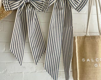 May include: Two large decorative bows with navy blue and white stripes hang on brass hooks against a white brick wall. The bows have long, flowing tails. A straw hat and a canvas tote bag are also visible.