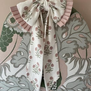 May include: A decorative bow made of white fabric with a floral pattern in pink and green. The bow is attached to a chair back with a floral and leaf pattern in shades of green, pink, and grey. The bow has pink striped trim.