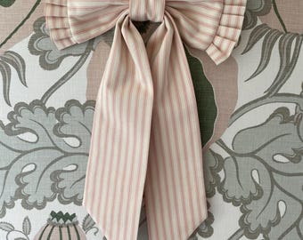 May include: A large, decorative bow made of striped fabric in cream and pale pink. The bow features pleated detailing on the sides and long, flowing tails. The bow is likely used for decoration or as an accessory.