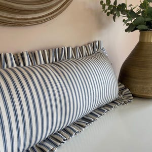 A long, white and blue striped cushion with a ruffled edge. The cushion is on a white surface.