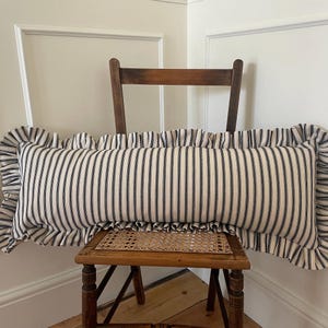 A long, white and black striped cushion with a ruffled edge. The cushion is sitting on a wooden chair with a woven seat.