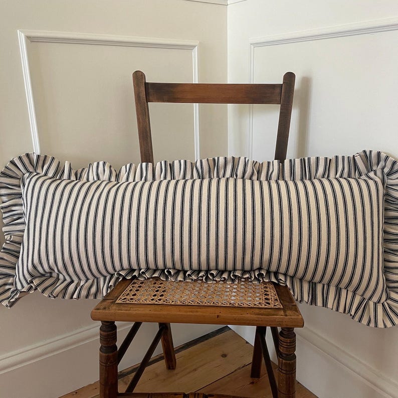 A long, white and black striped cushion with a ruffled edge. The cushion is sitting on a wooden chair with a woven seat.