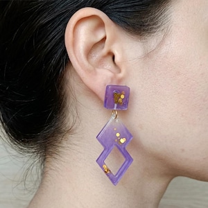 Peut inclure: Boucles d'oreilles en résine violette avec des paillettes dorées. Les boucles d'oreilles présentent un clou carré et un pendant en forme de losange. Les bijoux sont attachés avec un fil métallique doré. Les boucles d'oreilles sont portées.