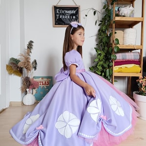 Puede incluir: Un vestido largo de color lavanda con mangas cortas abullonadas, una falda amplia con grandes aplicaciones florales blancas y una enagua de tul rosa. El vestido incluye una diadema lavanda a juego con un lazo rosa y una cinta en la cintura.