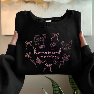 May include: Black crewneck sweatshirt with a pink graphic. The design includes a cow, chicken, vegetables, and the words "homestead mama". The sweatshirt is held up by two hands.
