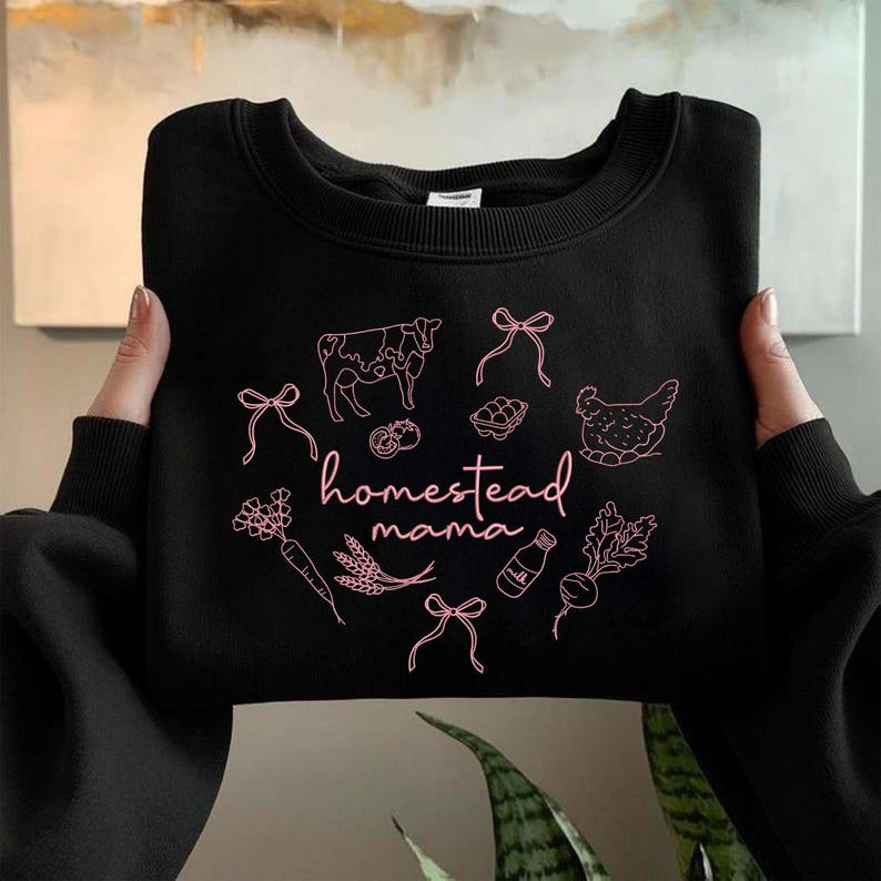 May include: Black crewneck sweatshirt with a pink graphic. The design includes a cow, chicken, vegetables, and the words "homestead mama". The sweatshirt is held up by two hands.