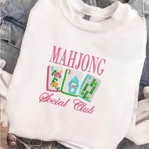 Może przedstawiać: Biała bluza z haftowanym r&oacute;żowym napisem "MAHJONG Social Club". Poniżej trzy haftowane kafelki do Mahjonga w kolorze r&oacute;żowym, zielonym i niebieskim. Bluza wykonana jest z miękkiego materiału.