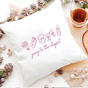 May include: White square pillow with a pink line drawing of wedding-related icons, including a bouquet, shoes, ring, champagne, and glasses. The phrase "going to the chapel" is written in cursive below the icons.