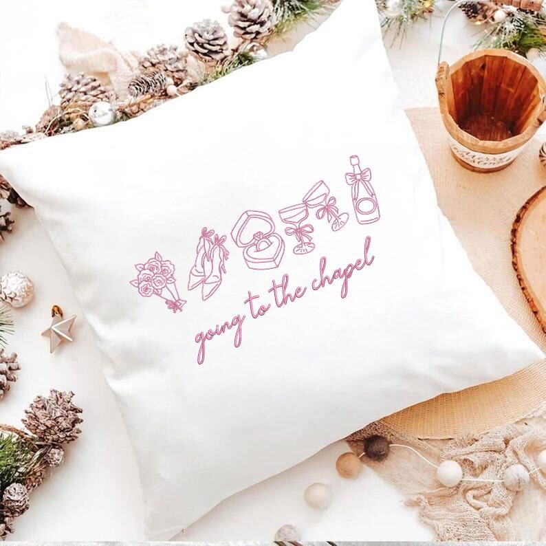 May include: White square pillow with a pink line drawing of wedding-related icons, including a bouquet, shoes, ring, champagne, and glasses. The phrase "going to the chapel" is written in cursive below the icons.